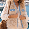 Gilet sans manches en fausse fourrure écrue avec poches et finitions en jean bleu, porté sur une robe bohème blanche.